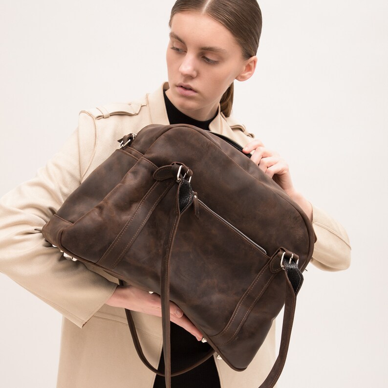 May include: A brown leather satchel bag with a black strap and silver hardware. The bag has a zippered pocket on the front and a flap closure.