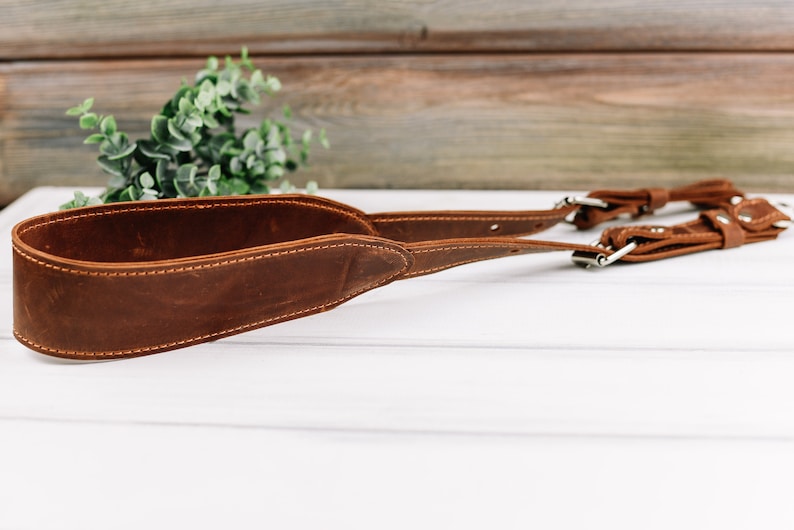 Peut inclure: Une sangle de cam&eacute;ra en cuir marron avec des coutures et des accessoires argent&eacute;s.