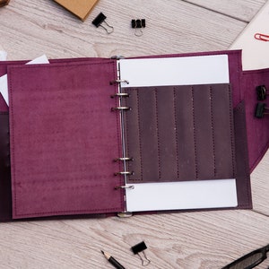 May include: A burgundy leather bound journal with a ring binder and a pocket on the front cover. The journal is open to a blank page with a pocket on the right side.