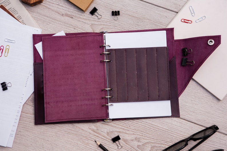 May include: A burgundy leather bound journal with a ring binder and a pocket on the front cover. The journal is open to a blank page with a pocket on the right side.