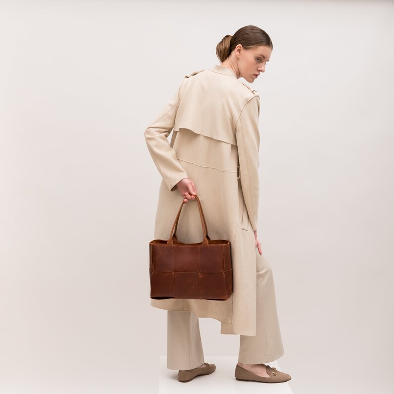 May include: A woman wearing a beige trench coat and beige pants holds a brown leather tote bag with a woven design.