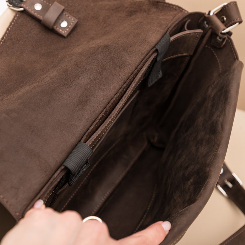 May include: A brown leather messenger bag with a flap closure and a black strap. The bag has a zippered pocket on the front and a large main compartment.