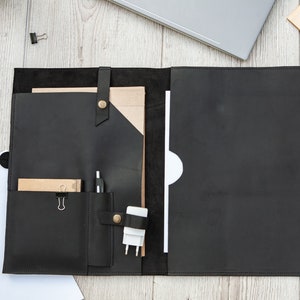 May include: Black leather portfolio with a flap closure, multiple pockets, and a strap. The portfolio is open and contains a notepad, pen, and a phone charger.