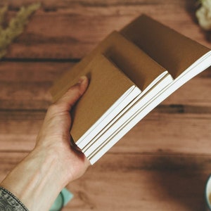 Puede incluir: Una mano sostiene una pila de tres cuadernos marrones con p&aacute;ginas blancas. Los cuadernos est&aacute;n sobre una superficie de madera.