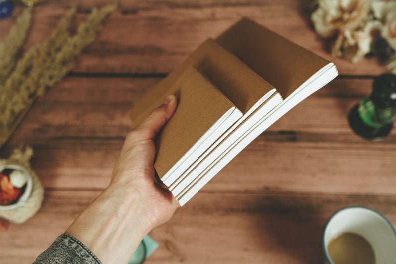 Puede incluir: Una mano sostiene una pila de tres cuadernos marrones con p&aacute;ginas blancas. Los cuadernos est&aacute;n sobre una superficie de madera.