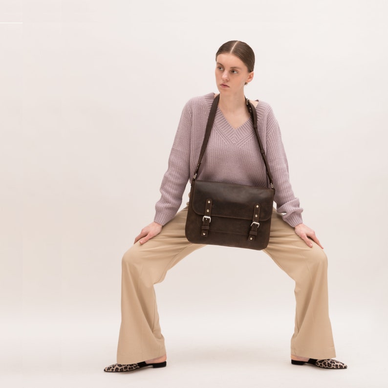 May include: A woman wearing a light purple sweater and beige pants is holding a brown leather satchel bag with buckles. The bag is worn over the shoulder and has a long strap.
