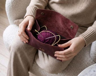 May include: A deep burgundy suede leather case with a fold-over flap, magnetic snap closures, and the embossed letters AB on the front. The interior contains a ball of dark purple yarn and several metal knitting needles.