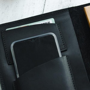 May include: Black leather notebook cover with a phone and card holder. The cover has a stitched edge and a brown notebook inside.