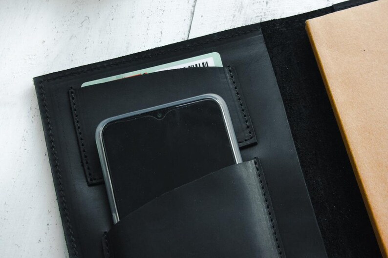May include: Black leather notebook cover with a phone and card holder. The cover has a stitched edge and a brown notebook inside.