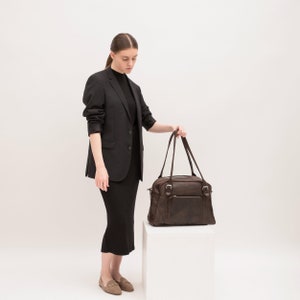 May include: A woman wearing a black blazer and skirt stands in front of a white background. She is holding a brown leather travel bag with a zippered pocket and buckles.