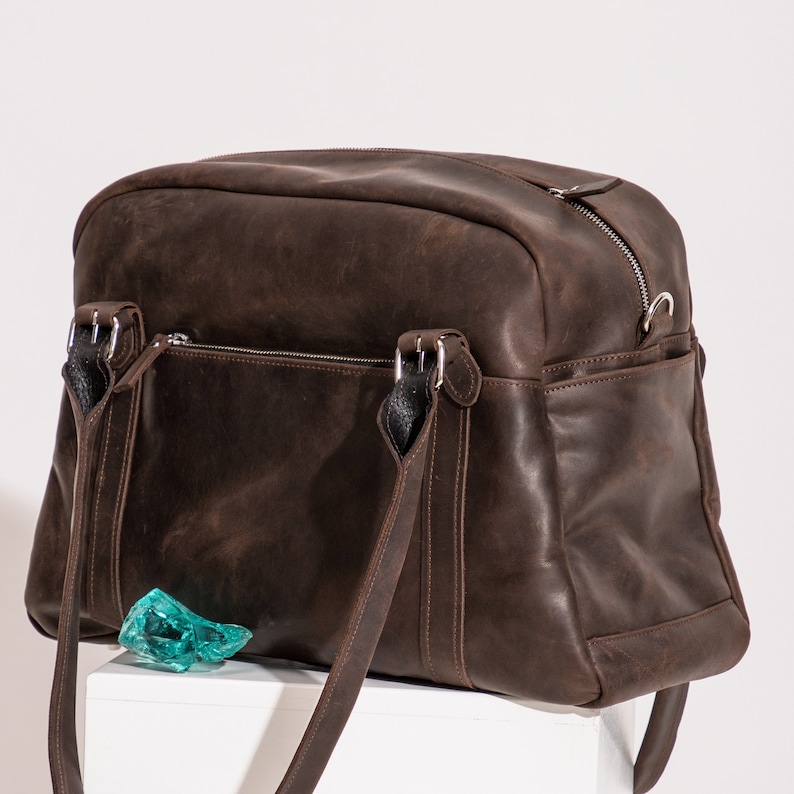May include: A brown leather duffel bag with two top handles and a zipper closure. The bag has a front pocket and a side pocket.