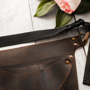 May include: A brown leather tool belt with multiple pockets and a black adjustable strap. The belt has brass rivets and a metal buckle.