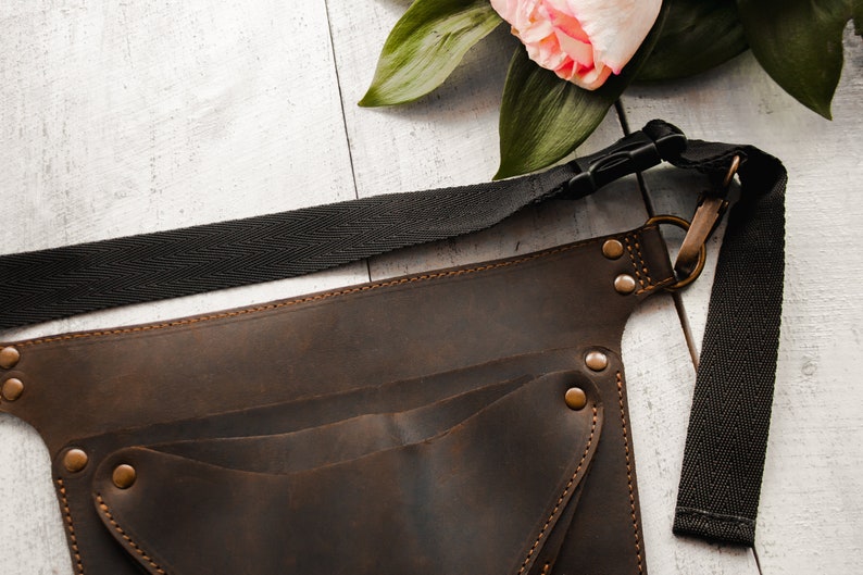 May include: A brown leather tool belt with multiple pockets and a black adjustable strap. The belt has brass rivets and a metal buckle.