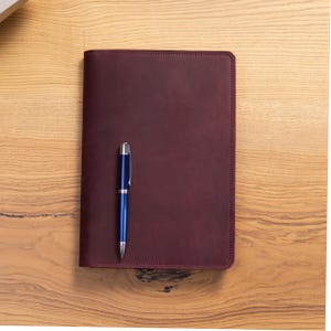 Puede incluir: Un diario de cuero burdeos con un bol&iacute;grafo azul encima. El diario es rectangular y tiene un borde cosido. El bol&iacute;grafo es plateado y azul. El diario y el bol&iacute;grafo est&aacute;n sobre una superficie de madera.