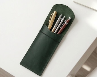 May include: A dark green leather pen case with a rounded top flap, secured by a thin black elastic band. The case holds four writing instruments: a gold-toned hexagonal pen, a silver metal pen, a thin silver mechanical pencil, and a maroon pen.