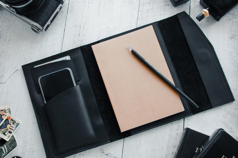 May include: A black leather notebook cover with a built-in pocket for a phone and cards. The cover is open to reveal a blank notebook and a black pen.