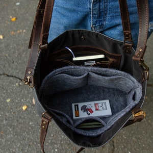 May include: A grey canvas tote bag with brown leather straps and a grey felt interior. The bag has multiple pockets and compartments, including a zipped pocket. The bag is open and the interior is visible, showing a grey felt liner with multiple pockets.