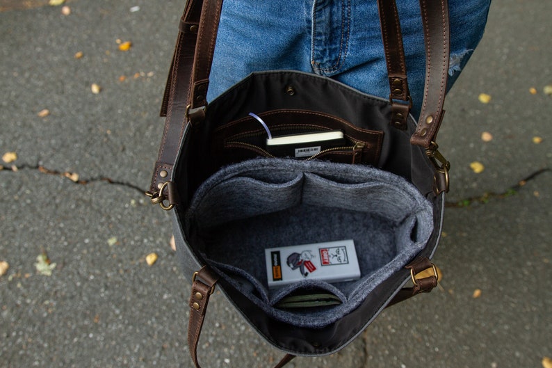 May include: A grey canvas tote bag with brown leather straps and a grey felt interior. The bag has multiple pockets and compartments, including a zipped pocket. The bag is open and the interior is visible, showing a grey felt liner with multiple pockets.