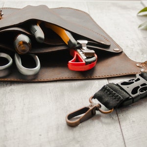 May include: A brown leather tool belt with a black strap and a buckle. The belt has multiple pockets and is filled with various tools, including a pair of scissors, a utility knife, and a wire stripper.