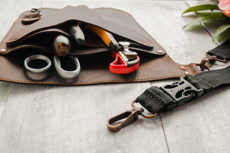 May include: A brown leather tool belt with a black strap and a buckle. The belt has multiple pockets and is filled with various tools, including a pair of scissors, a utility knife, and a wire stripper.