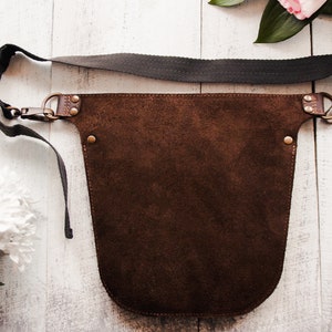 May include: A brown leather apron with a dark gray adjustable strap. The apron has a wide, rounded bottom and is secured with brass buckles and rivets.