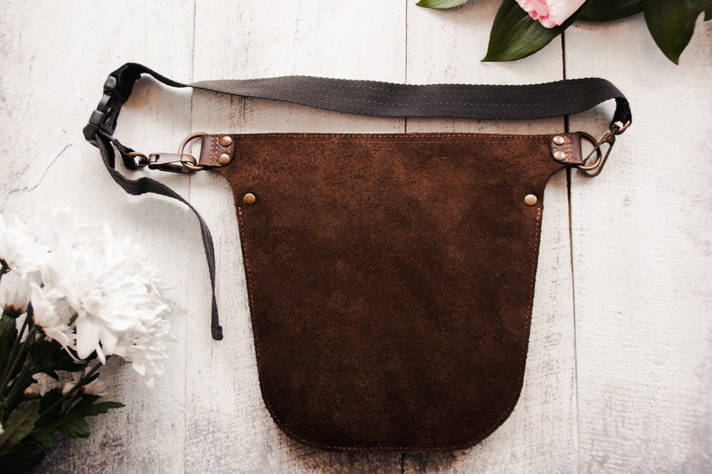 May include: A brown leather apron with a dark gray adjustable strap. The apron has a wide, rounded bottom and is secured with brass buckles and rivets.
