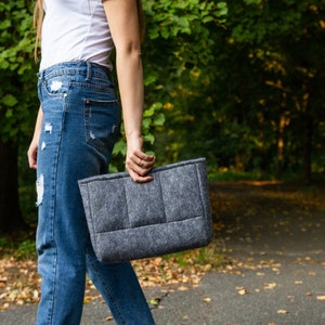 May include: A person wearing blue jeans and a white t-shirt is holding a grey felt tote bag.