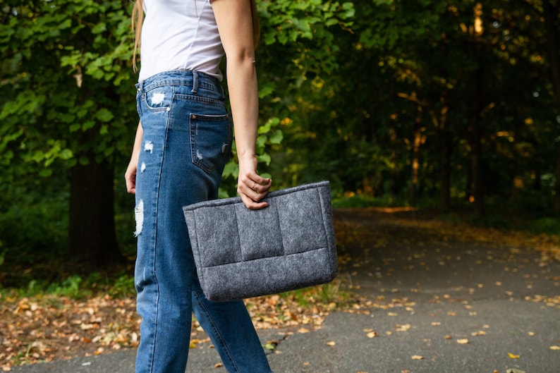 May include: A person wearing blue jeans and a white t-shirt is holding a grey felt tote bag.