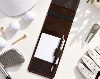 May include: A dark brown leather golf scorecard holder featuring a grid-lined notepad and a black and gold pen. The holder includes a black elastic strap and a leather pen loop. It is surrounded by golf balls, tees, a club, a glove, and a tool.