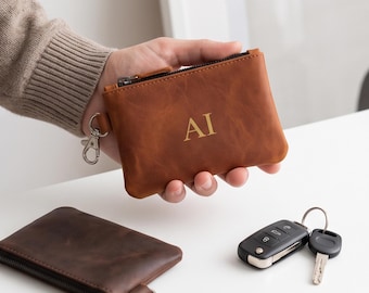 May include: A tan leather zippered pouch featuring the embossed letters AI on the front. The pouch includes a silver-toned metal lobster clasp attached to a side tab. A second, darker brown leather pouch and a car key fob are also visible.