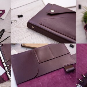 May include: A burgundy leather bound journal with a pocket on the front cover. The journal has a ring binder system and a flap that closes with a button. The journal is open to reveal a blank page and a pocket on the inside cover.