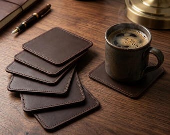 May include: A set of square dark brown leather coasters with rounded corners and visible stitching along the edges. One coaster supports a ceramic mug filled with dark coffee, while the remaining coasters are stacked in a neat pile.