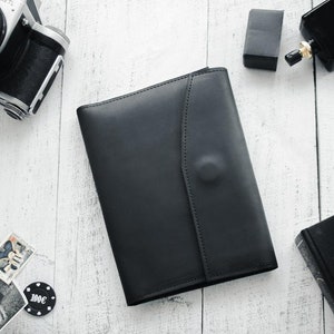 May include: A black leather portfolio with a flap closure. The portfolio is on a white wooden surface with other items, including a camera, a bottle of perfume, and a stack of books.