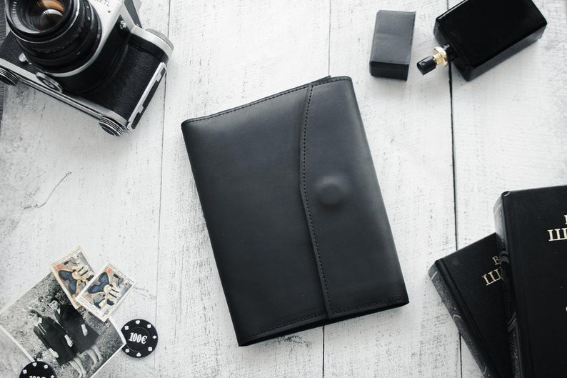 May include: A black leather portfolio with a flap closure. The portfolio is on a white wooden surface with other items, including a camera, a bottle of perfume, and a stack of books.