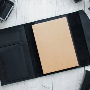 May include: A black leather portfolio with a brown paper insert and two pockets on the front. The portfolio is open and the paper insert is visible.