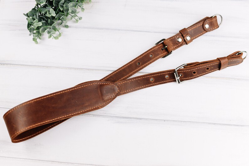 Peut inclure: Une sangle de cam&eacute;ra en cuir marron avec deux boucles r&eacute;glables et un anneau m&eacute;tallique. La sangle est con&ccedil;ue pour porter un appareil photo autour du cou ou de l'&eacute;paule.