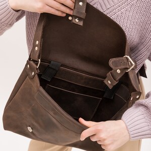 May include: A brown leather messenger bag with a flap closure and a buckle. The bag has a large main compartment and a smaller pocket on the front. The bag is open and the inside is visible.