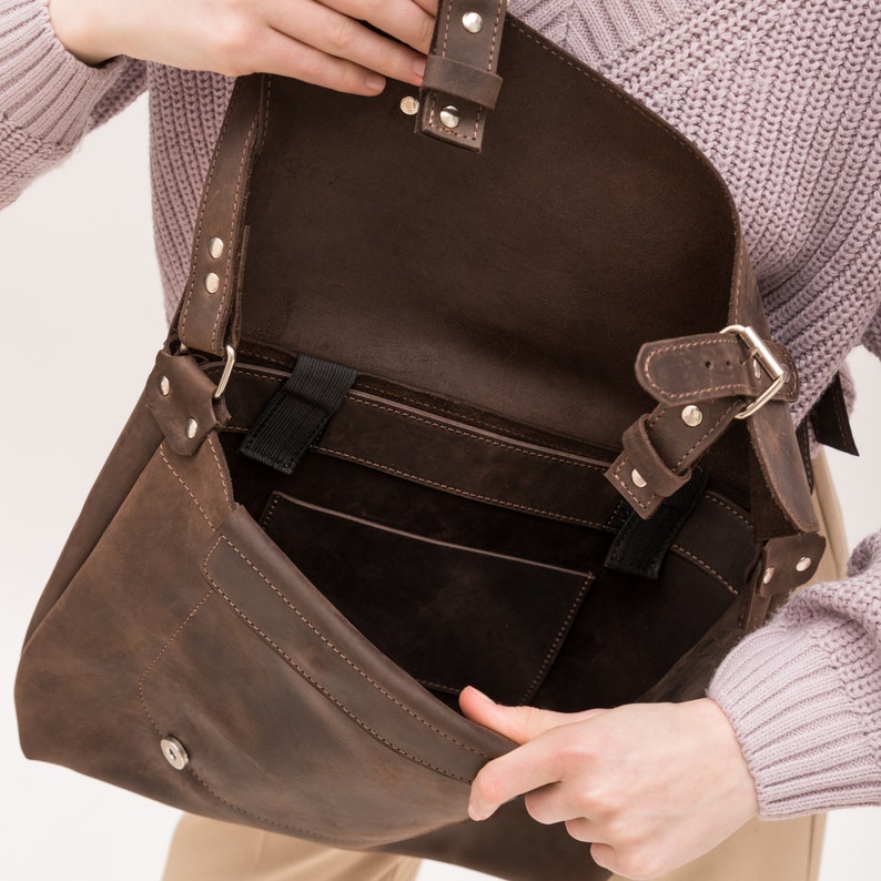 May include: A brown leather messenger bag with a flap closure and a buckle. The bag has a large main compartment and a smaller pocket on the front. The bag is open and the inside is visible.
