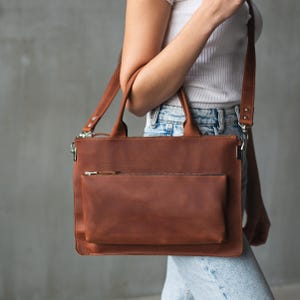 May include: A brown leather satchel with a shoulder strap and top handles. The bag has a front zippered pocket and a main compartment with a zipper closure. The bag is a medium size, suitable for carrying a laptop or other essentials. The bag is made of a smooth, high-quality leather.