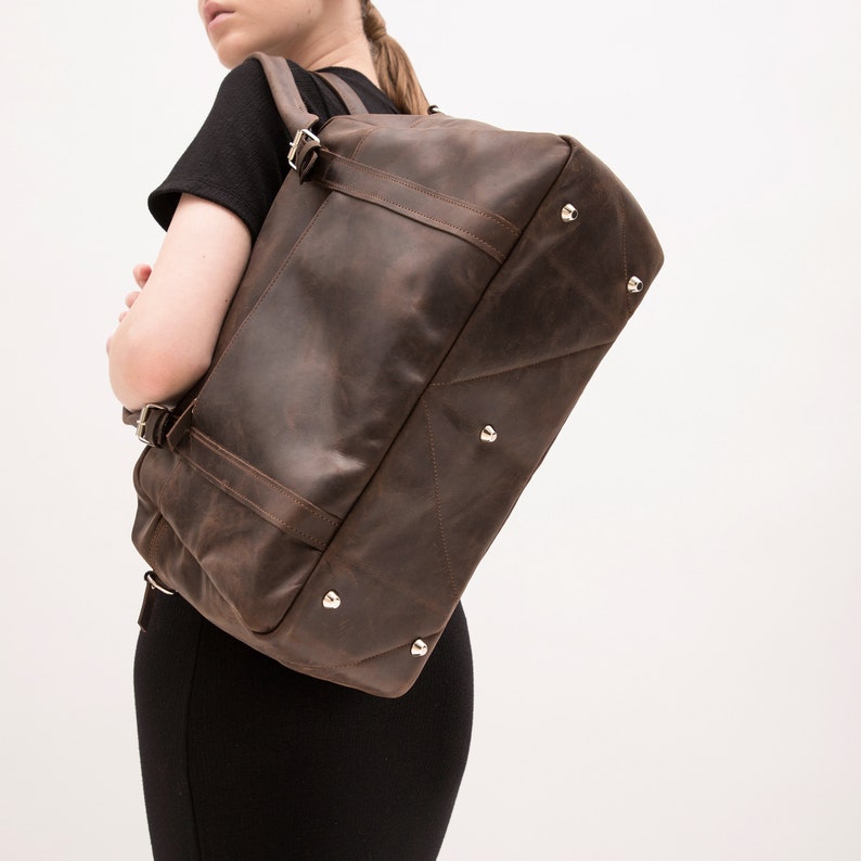 May include: A brown leather duffel bag with silver hardware and adjustable straps. The bag has a distressed look and is worn over the shoulder.