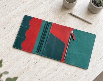 May include: A leather notebook cover featuring a color-blocked design in deep teal and vibrant red. The interior includes multiple card slots with scalloped edges, three elastic bands for securing inserts, a large slip pocket, and a zippered pouch.