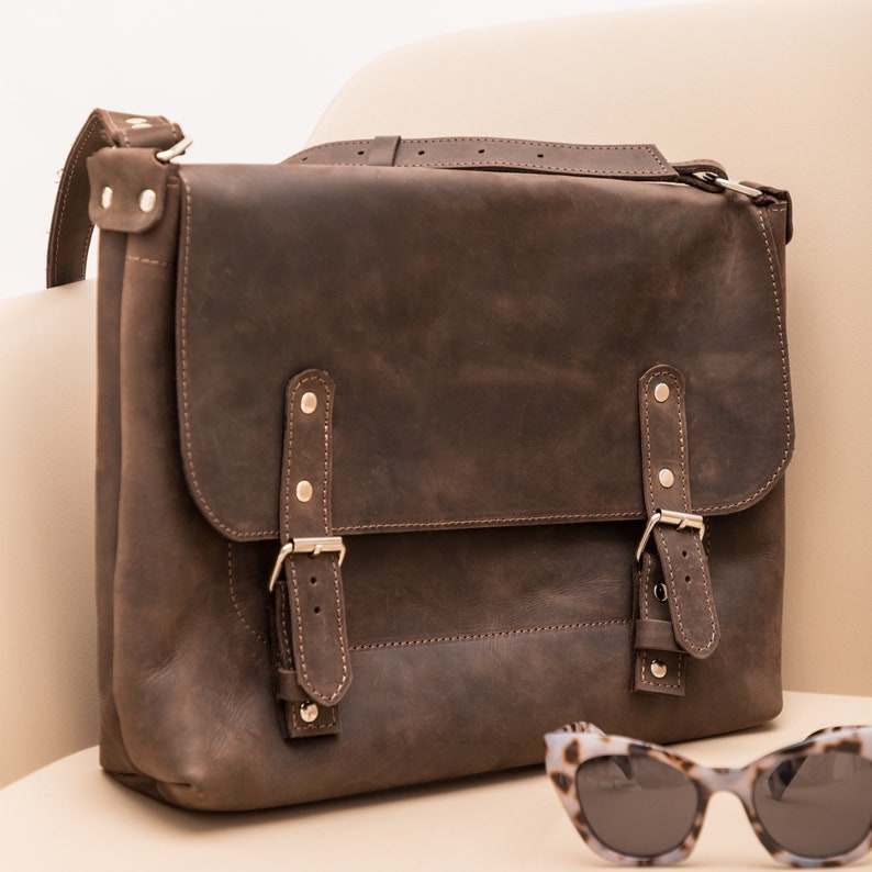 May include: A brown leather messenger bag with two buckles and a long strap. The bag is sitting on a light brown surface next to a pair of tortoiseshell sunglasses.