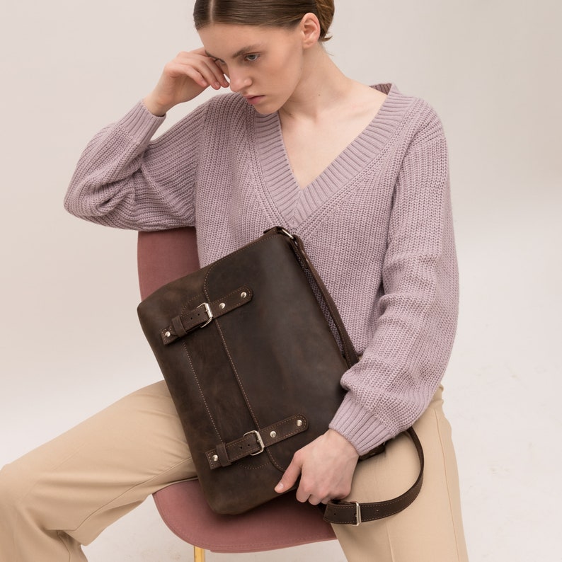 May include: A brown leather messenger bag with a buckle closure and a long adjustable strap. The bag is worn by a person wearing a light purple sweater and beige pants.