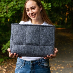 May include: A grey felt tote bag with a quilted design. The bag is held in front of a person wearing a white shirt and blue jeans.