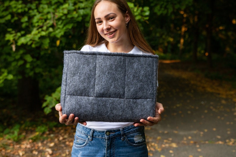 May include: A grey felt tote bag with a quilted design. The bag is held in front of a person wearing a white shirt and blue jeans.