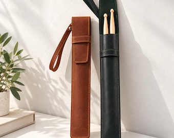 May include: Two tall, slender leather cases for drumsticks, one in a rich brown and the other in a deep black. Each case features a matching leather wrist strap, a secure flap closure, and visible stitching. The black case holds two wooden drumsticks.