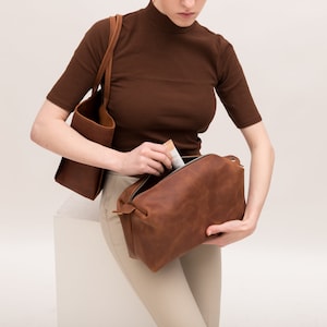 May include: A brown leather tote bag with a brown leather zippered pouch. The pouch is open and a person is putting something inside.