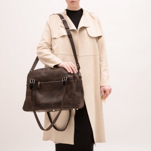 May include: A brown leather satchel bag with a long strap. The bag has two top handles and a zipper closure. The bag is being held by a person wearing a beige coat.