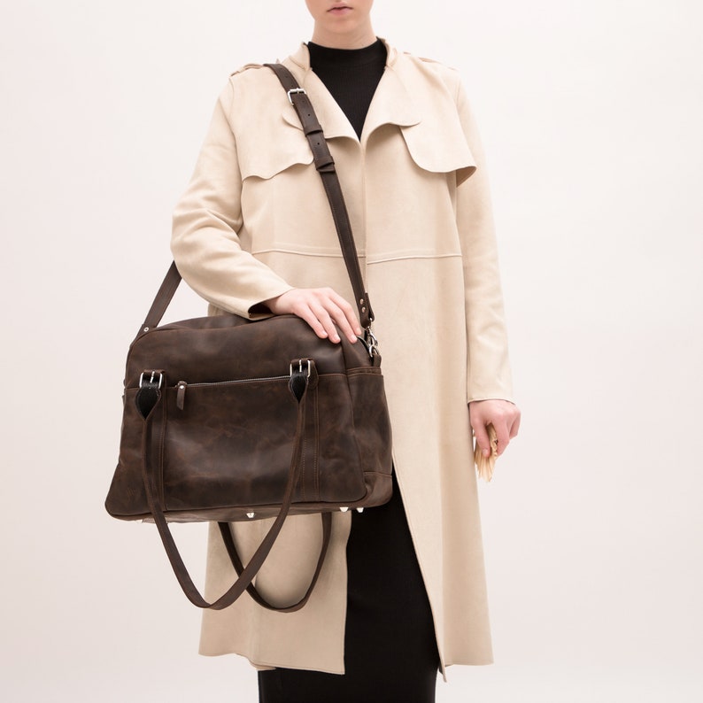 May include: A brown leather satchel bag with a long strap. The bag has two top handles and a zipper closure. The bag is being held by a person wearing a beige coat.