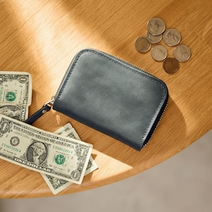 Könnte beinhalten: Dunkelgraue Ledergeldbörse mit Reißverschluss, kleiner Handschlaufe und mehreren US-Dollar-Scheinen und Münzen auf einer Holzoberfläche. Die Geldbörse ist rechteckig.